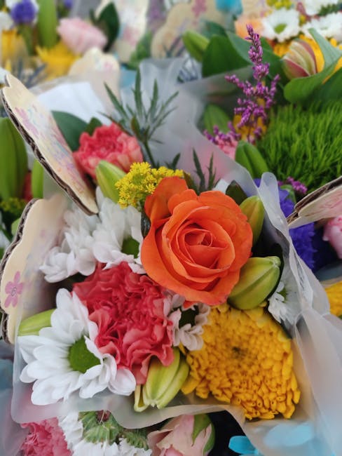 A vibrant floral arrangement featuring a central orange rose surrounded by white daisies, pink carnations, yellow chrysanthemums, and purple lavender, all arranged with green foliage and wrapped in wh