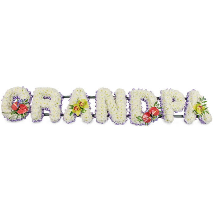 This long, horizontal memorial arrangement spells the word "GRANDPA" in bold capital letters, each one sculpted from a mass of fresh, snowy white chrysanthemums. The flowers are packed closely together so their delicate petals form a plush, velvety surface, catching gentle highlights and shadows that emphasise the contours of every letter. The tribute is photographed against a simple white background, allowing the shapes and textures to stand out without distraction. Along the base of each letter, a narrow ruffle of pale lavender ribbon traces a soft outline, the lilac hue bringing a quiet, cool contrast that frames the word with understated elegance. Dark green metal or wooden bars support the letters from beneath, mostly concealed yet essential for keeping the tribute secure during transport across Northolt for a funeral or cremation service. Carefully placed accent clusters punctuate the design: both the "G" and the "D" are adorned with rich sprays of deep red anthuriums, their smooth, heart-shaped, almost waxy spathes glistening under the light, each with a vivid yellow spadix rising from the centre. These bold blooms sit amongst glossy dark green foliage, symbolising heartfelt love and remembrance. On the first and last "A," sprays of yellow-green cymbidium orchids rest elegantly along the chrysanthemum base, their arched stems and patterned throats suggesting refinement and lasting affection. Smaller leaves and filler flowers extend naturally from each cluster, creating gentle movement across the arrangement while preserving its clean, ordered structure. The result feels calm and sincere, a thoughtfully designed "GRANDPA" tribute that families in Northolt might choose to place by the coffin, at the front of the service, or on the grave as a final, floral thank-you to a much-loved grandfather.